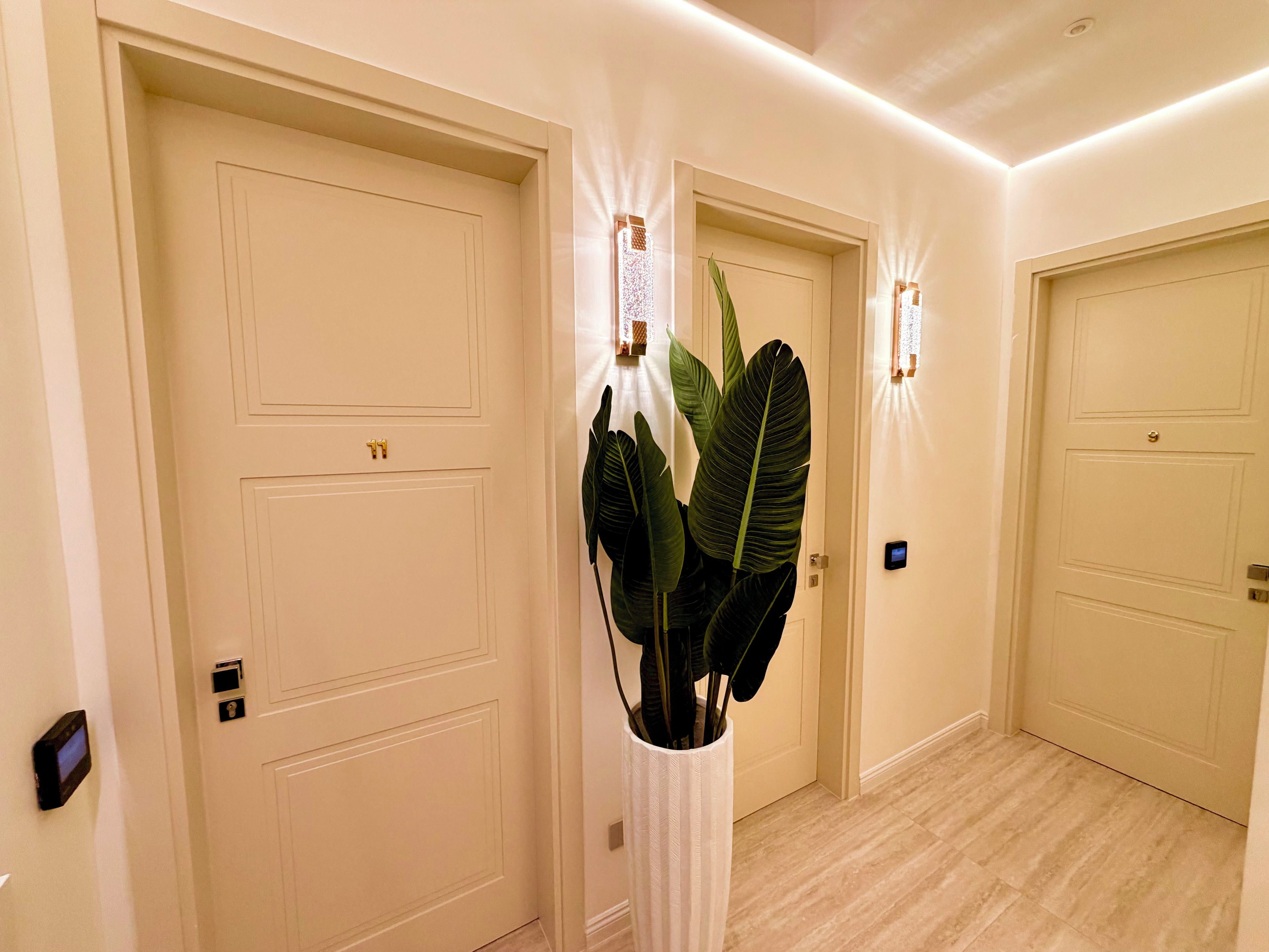 Corredor com três portas fechadas, um vaso de planta e um luminário fixado na parede.