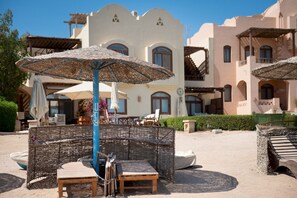 Outdoor seating with woven umbrellas, a poolside area, and a building with arched windows and balconies. - Vesta Luxury Apartment -1br - New Nubia (El Gouna)
