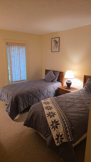 Ein Schlafzimmer mit zwei Betten, einem Nachttisch mit Lampe, einem Fenster mit Jalousien und einem Bilderrahmen an der Wand.