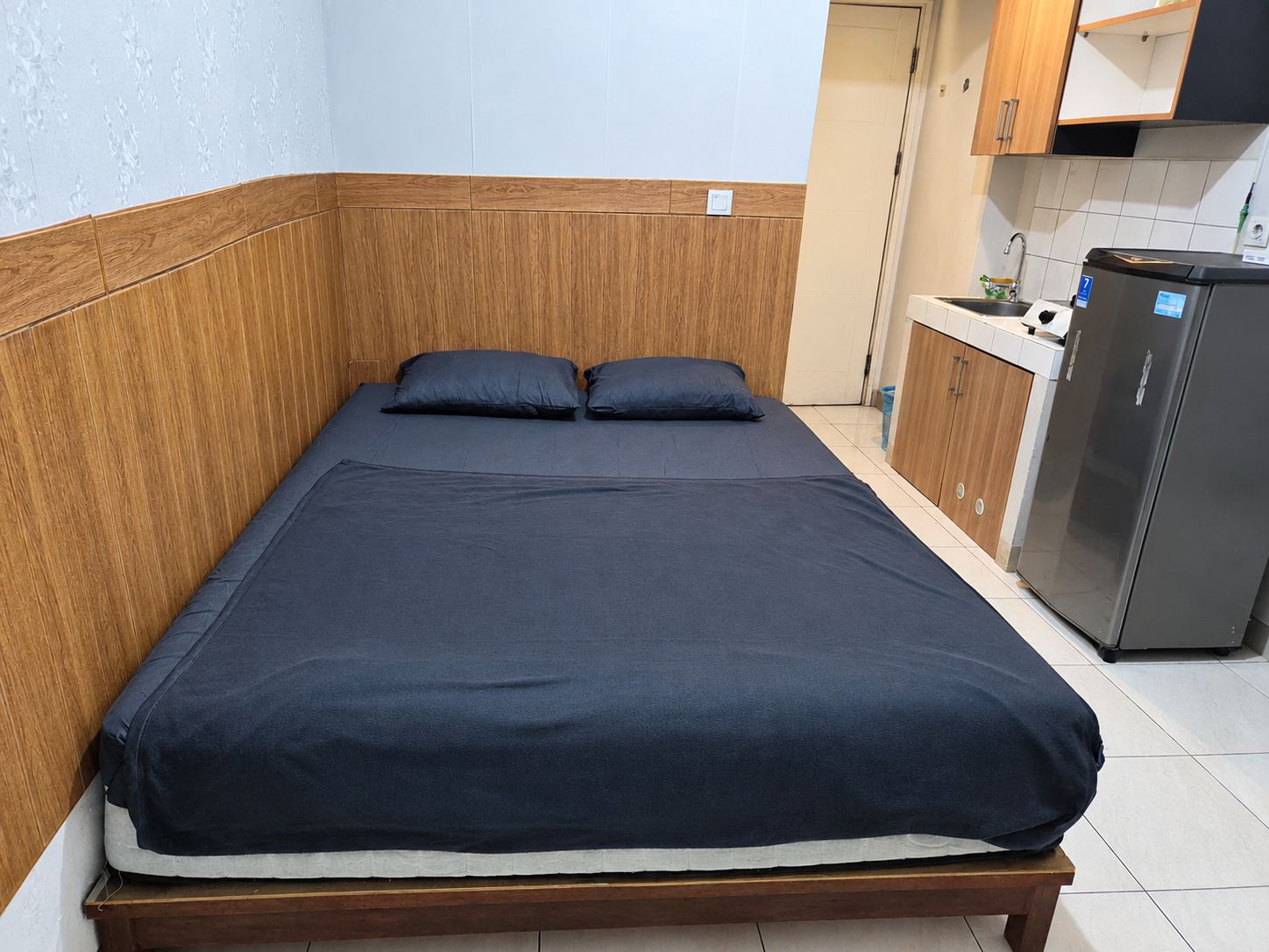 A single bed with a dark blue mattress and two pillows, a wooden headboard, and a kitchenette with a refrigerator and cabinets.