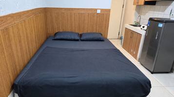 A single bed with a dark blue mattress and two pillows, a wooden headboard, and a kitchenette with a refrigerator and cabinets.