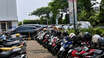 A row of parked motorcycles and cars in a parking lot with trees and a building in the background.