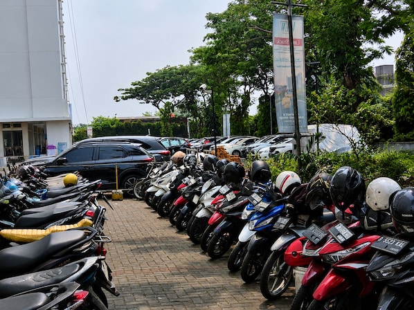 Una fila de motocicletas y automóviles estacionados en un estacionamiento, con árboles y un edificio al fondo.