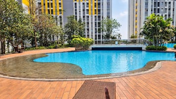 Piscina com deck de madeira e espreguiçadeiras, cercada por vegetação e prédios altos.