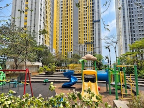 A playground with various play structures, surrounded by greenery and tall residential buildings in the background. - Nusantara Rooms Springlake By Reccoma (Bekasi)