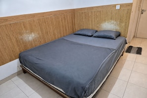 Un lit simple avec une couvre-lit grise et deux oreillers, une tête de lit en bois et un sol carrelé.