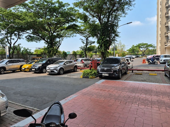 Un espace de stationnement où sont garées plusieurs voitures et une moto, entouré d’arbres et d’un bâtiment.