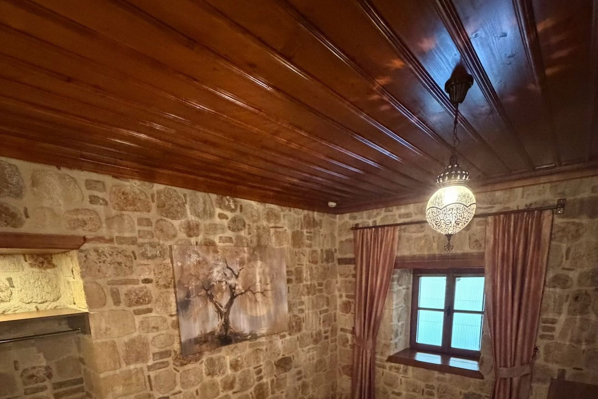 Ein Zimmer mit Steinwand, Holzboden, einem Bett mit Kissen, einem Nachttisch, einer Hängeleuchte und einem Fenster mit Vorhängen.