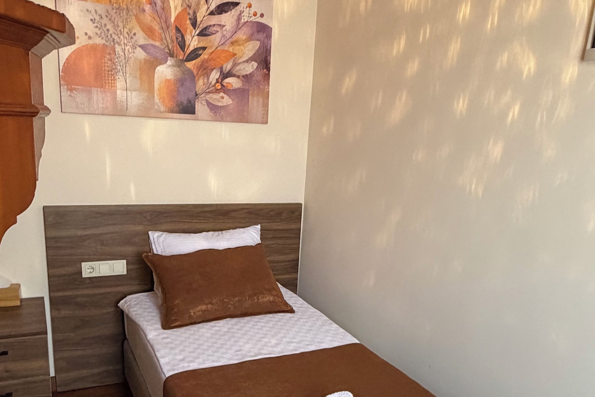 Ein Einzelbett mit brauner Kopfteil, einem weißen Kopfkissen und einer braunen Decke. Ein weißes Handtuch liegt gefaltet auf dem Bett.