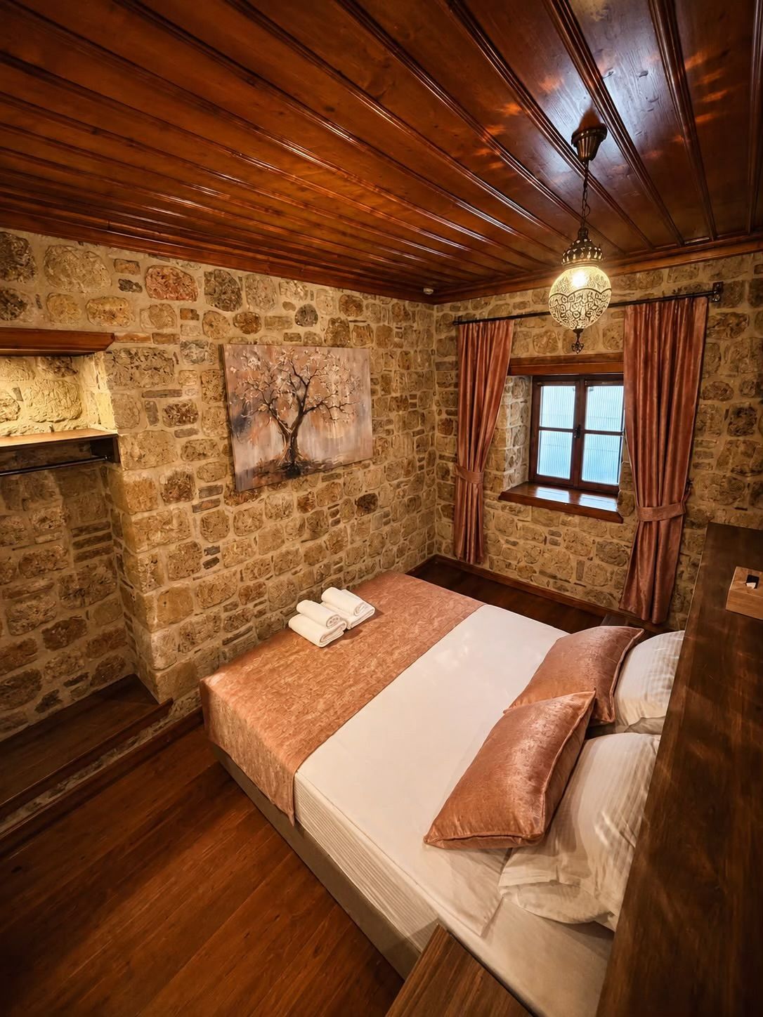 Un dormitorio con pared de piedra, una cama con edredón blanco y marrón, un cuadro de un árbol y una ventana con cortinas.