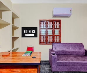 A hotel room with a wooden table, a purple sofa, a window with wooden shutters, and a sign indicating it is serviced by OYO.