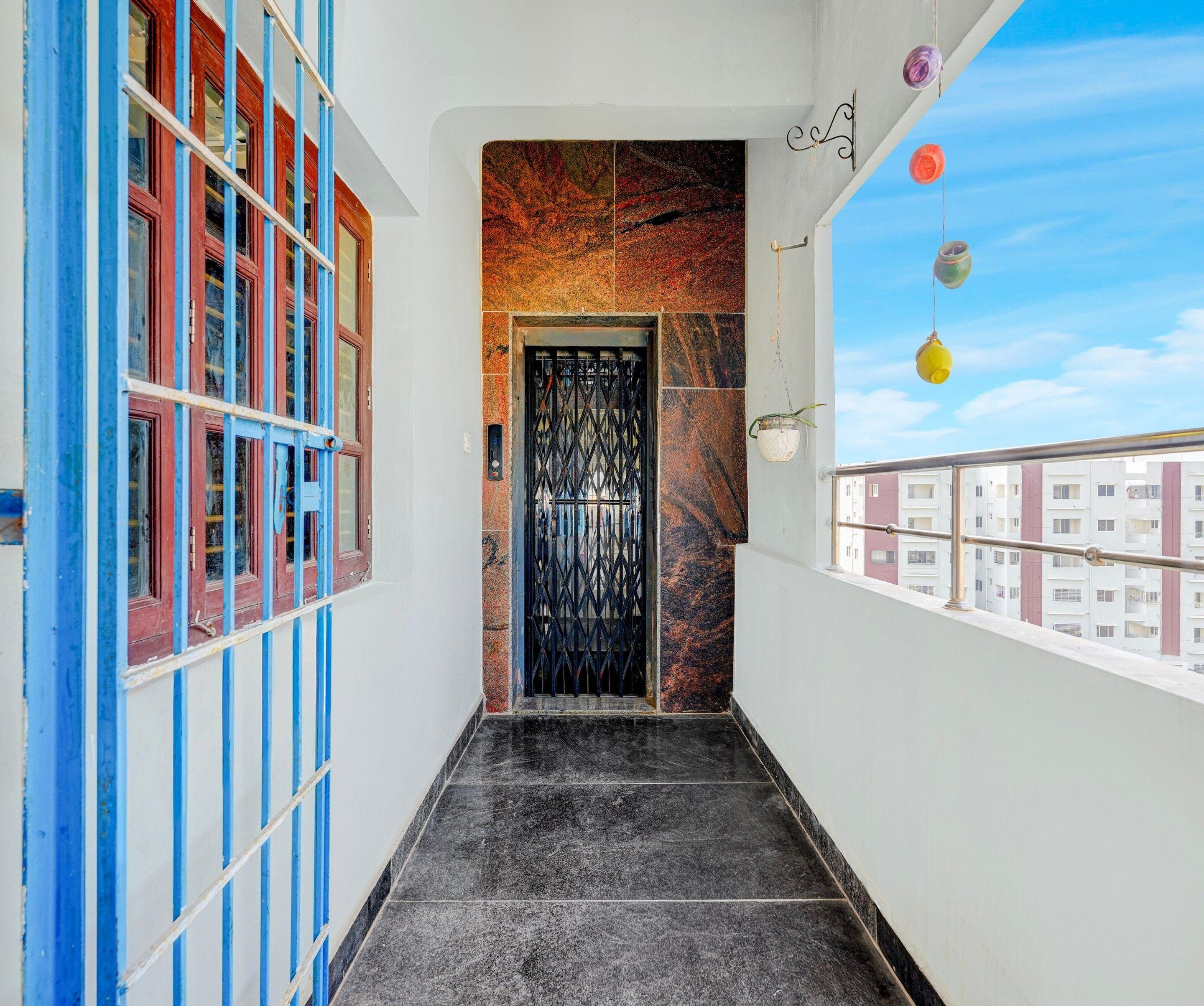 Un pasillo estrecho con una puerta de metal negro, una ventana azul y roja, y edificios a lo lejos.