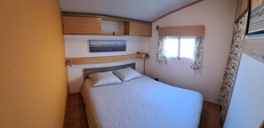 A bedroom with a bed, a headboard, a window with curtains, and a wall-mounted shelf with a picture. - 🌊 Fully equipped mobile home for 4 people – SIBLU Mimizan Plage   (Mimizan)