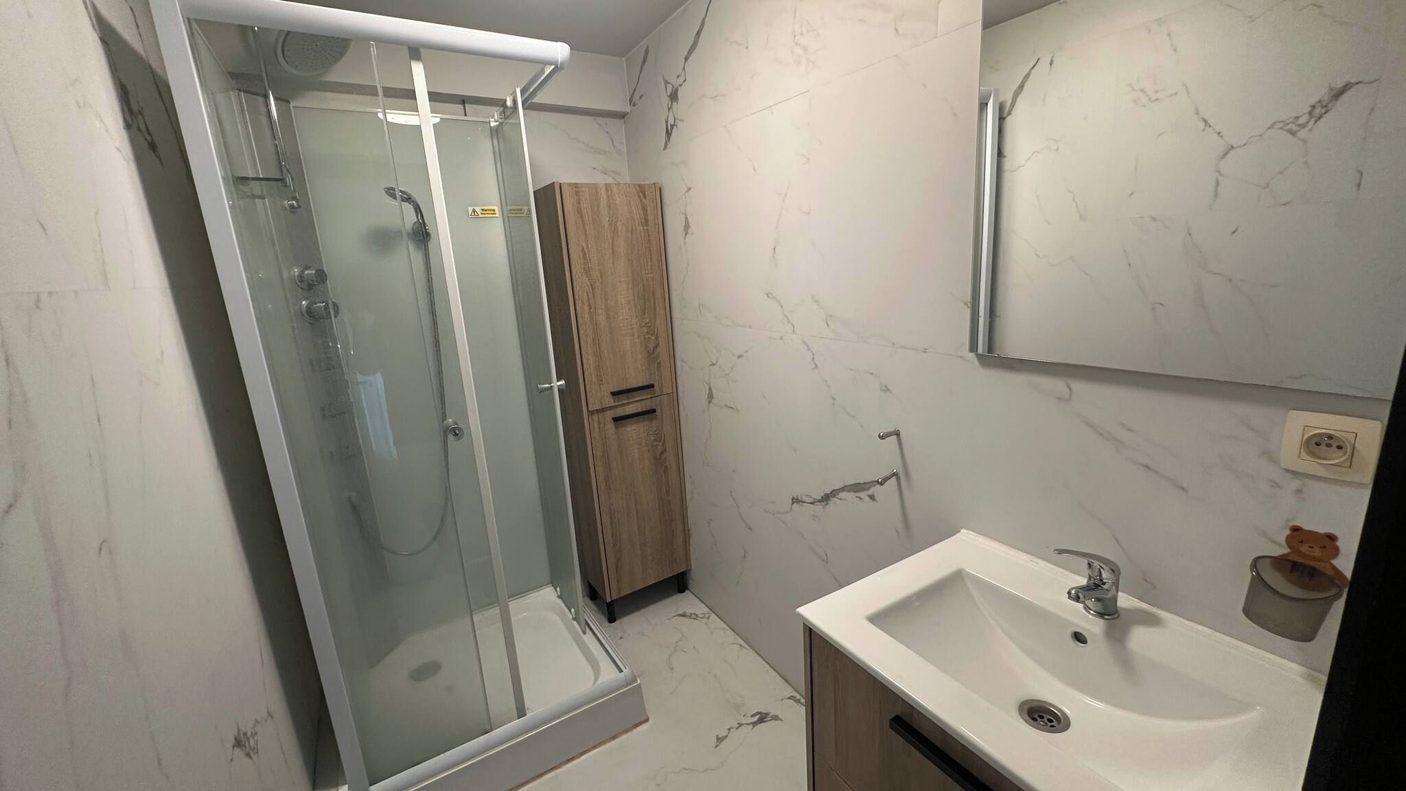 A bathroom with a glass-enclosed shower, a wooden cabinet, a sink with a mirror, and marble-patterned walls.