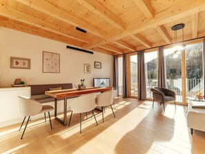 A spacious room with a wooden ceiling, a dining area with a long table and chairs, a sofa, and a large window offering a view of the outdoors. - Chalet #7 With 3 Bedrooms and Sauna (Oberwölz)