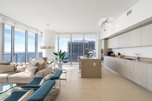 Un salon moderne comprenant un canapé, des fauteuils et un coin repas. On y trouve également une cuisine équipée de placards et d’un plan de travail. De grandes fenêtres offrent une vue sur la ville.