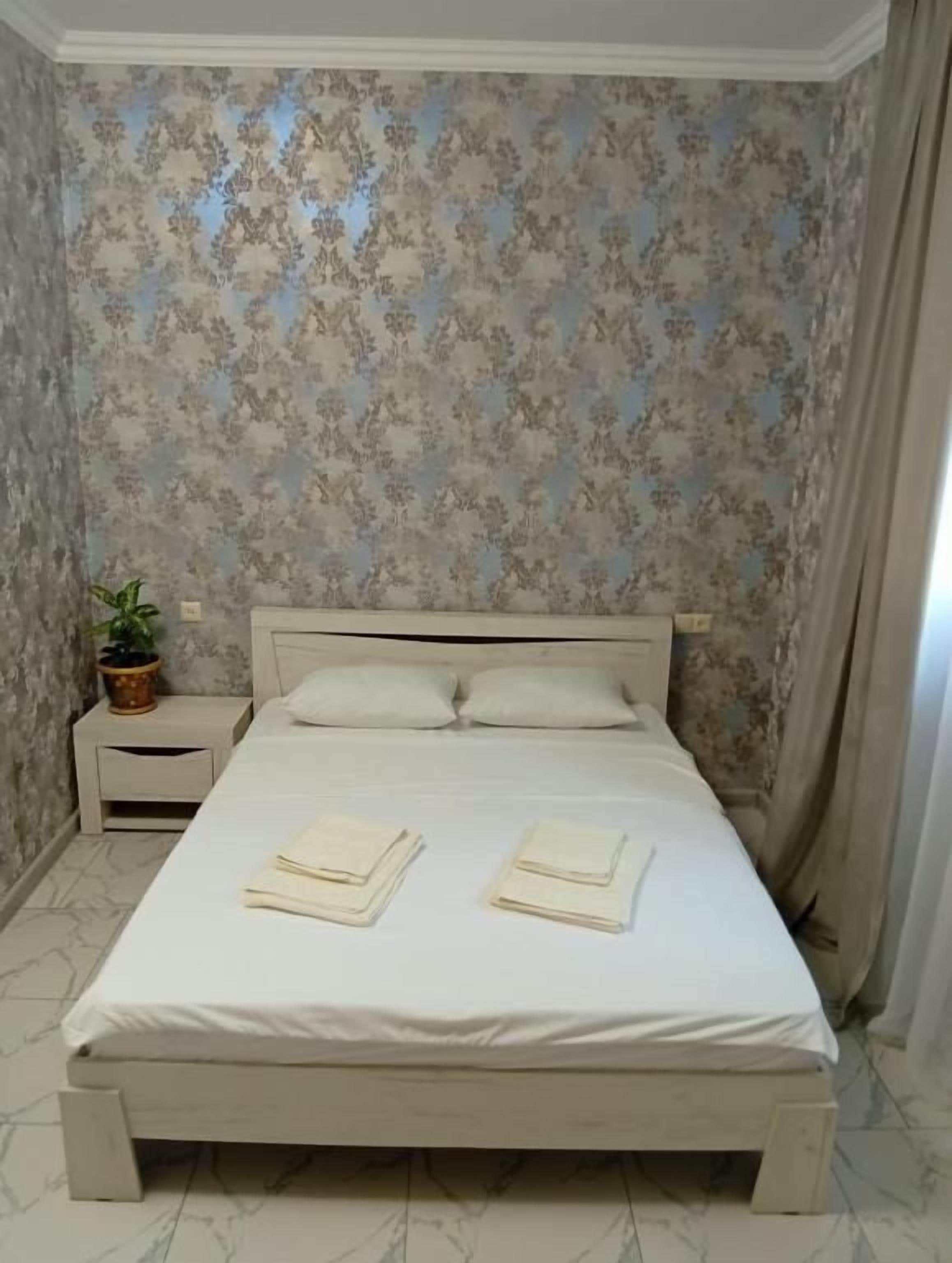 Una cama bien hecha con almohadas y toallas dobladas, una mesita de noche con una planta y una pared con papel tapiz floral.