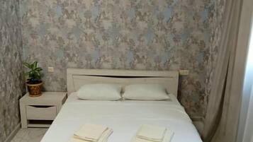 Una cama bien hecha con almohadas y toallas dobladas, una mesita de noche con una planta y una pared con papel tapiz floral.