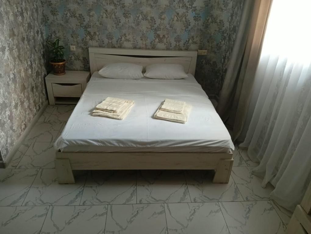 Un lit avec un matelas blanc et un cadre en bois, une table de chevet avec une plante, et une fenêtre avec des rideaux.