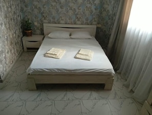 A bed with a white mattress and wooden frame, a nightstand with a plant, and a window with curtains. - Veni Vedi guesthouse  (Vedi)