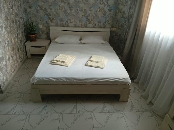 Una cama con colchón blanco y estructura de madera, una mesita de noche con una planta y una ventana con cortinas.