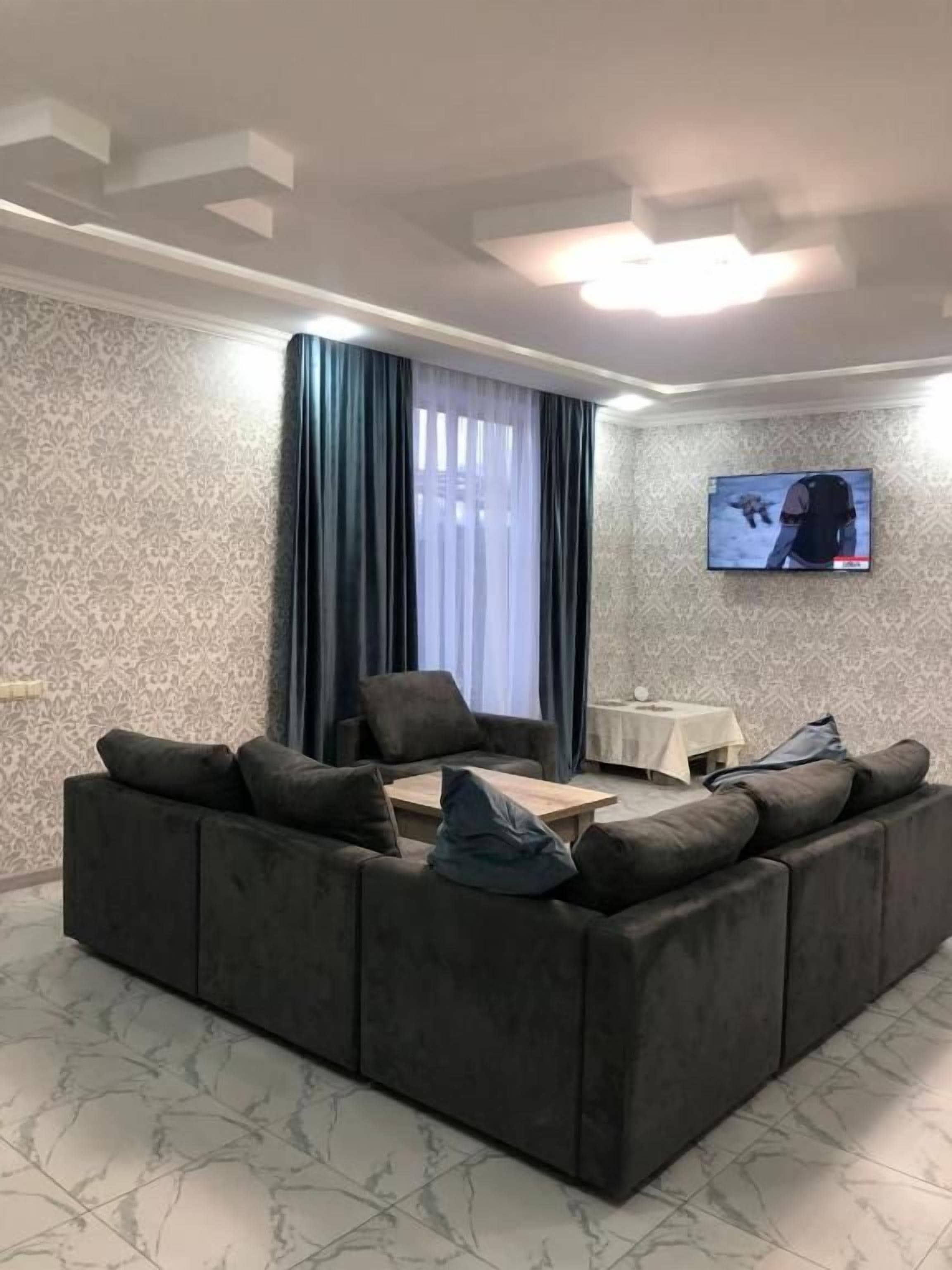Una sala de estar con un sofá seccional, un televisor de pantalla plana montado en la pared y una ventana con cortinas.