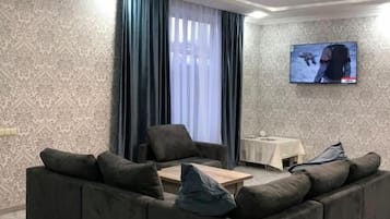 Una sala de estar con un sofá seccional, un televisor de pantalla plana montado en la pared y una ventana con cortinas.
