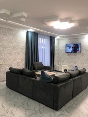 A living room with a sectional sofa, a flat-screen TV mounted on the wall, and a window with curtains. - Veni Vedi guesthouse  (Vedi)