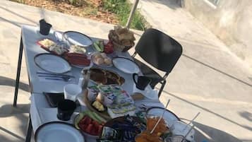 Mesa de comedor al aire libre con platos, cubiertos, una cesta de pan, un bol de fruta y bebidas.
