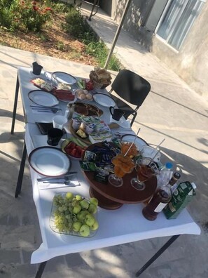 Mesa de comedor al aire libre con platos, cubiertos, una cesta de pan, un bol de fruta y bebidas.