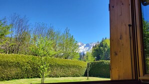Blick aus einem Fenster mit Holzrahmen auf einen gepflegten Garten mit Hecke, einem kleinen Baum und einem Bergkamm in der Ferne.