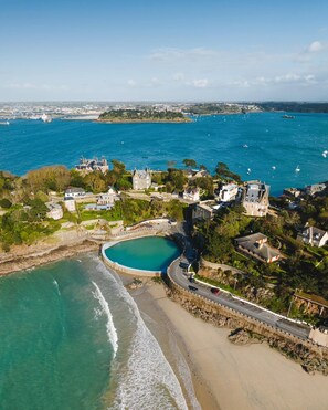 An aerial view of a coastal area with a large circular pool, beachfront properties, and a marina. - Vrbo Property (DINARD)
