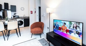 Ein modernes Wohnzimmer mit Flachbildfernseher, einem Essbereich mit Stühlen, einer Küche im Hintergrund und einem roten Sessel.