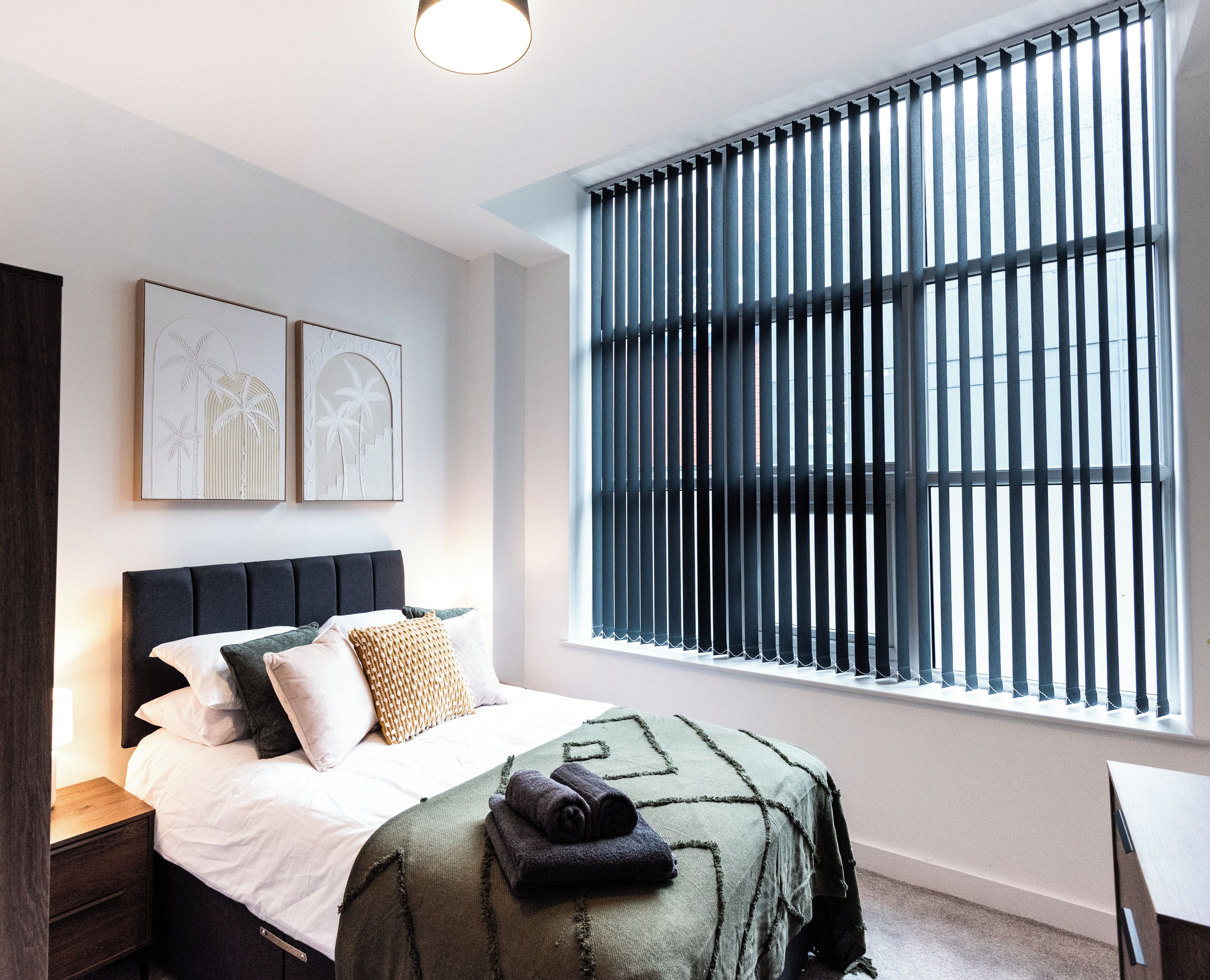 A bedroom with a bed, a nightstand, a window with blinds, and two wall-mounted artworks.