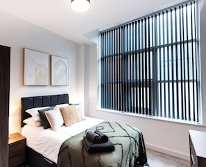A bedroom with a bed, a nightstand, a window with blinds, and two wall-mounted artworks. - Apex House by Guestz (Warrington)