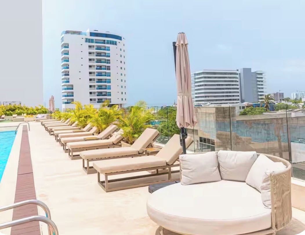 Un área de piscina en la azotea con sillones de descanso y un espacio de asientos con cojines, con vistas a un paisaje urbano.