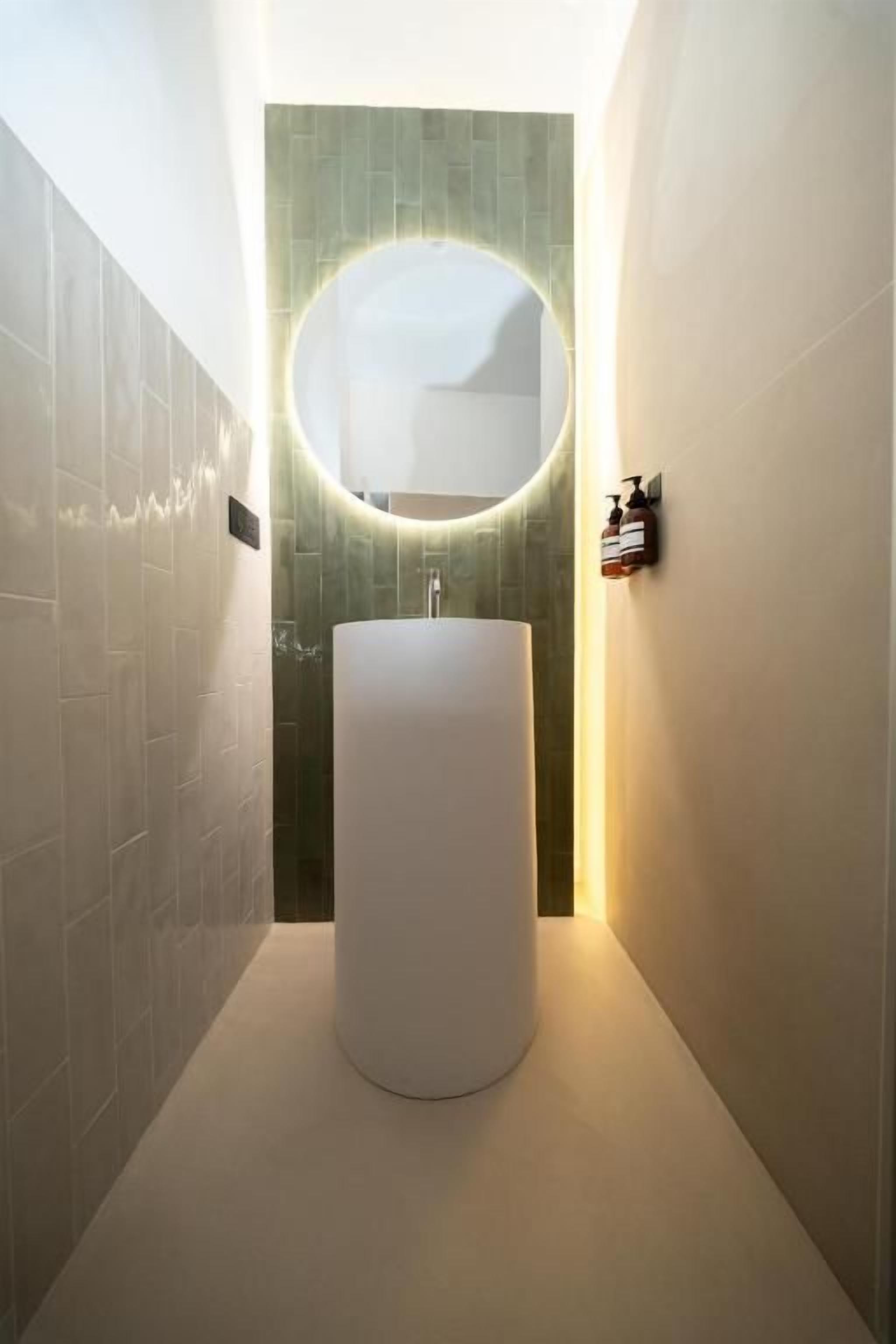 Une salle de bain moderne avec un miroir rond, un lavabo cylindrique et un mur recouvert de carreaux verts.