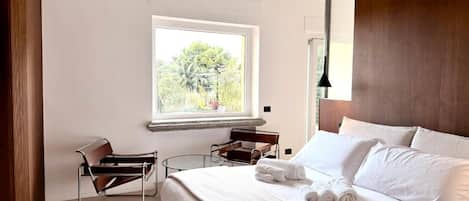 Habitación de hotel con cama, ventana con vista a la vegetación, una silla y una lámpara.
