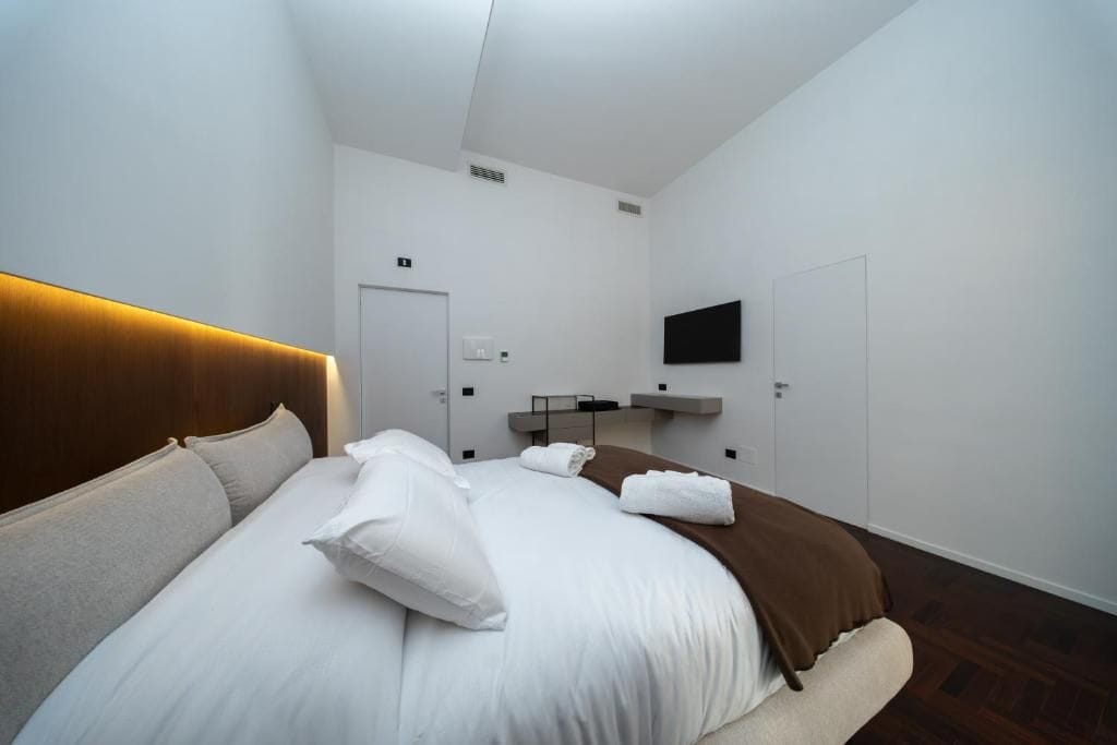 A hotel room with a bed, a flat-screen TV mounted on the wall, a door, and a small shelf with items.