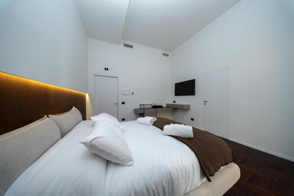 A hotel room with a bed, a flat-screen TV mounted on the wall, a door, and a small shelf with items.
