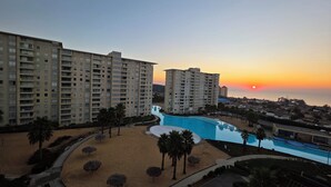 Een hoog hotel met zwembad en strand, omgeven door palmbomen en een uitzicht op de zonsondergang.