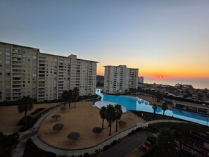 Een hoog hotel met zwembad en strand, omgeven door palmbomen en een uitzicht op de zonsondergang.