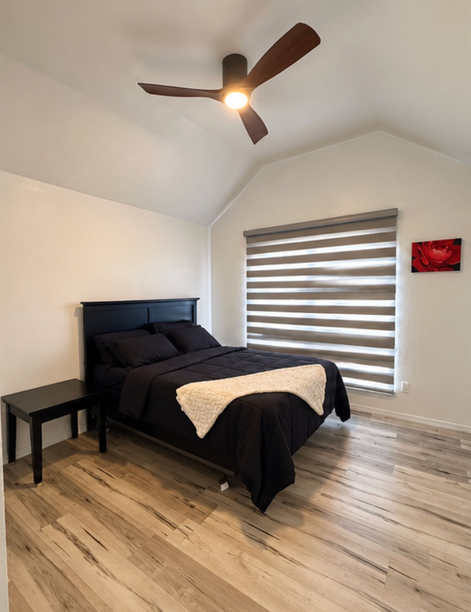 Une chambre à coucher comprenant un lit, une table de chevet, un ventilateur de plafond et une fenêtre avec des stores.