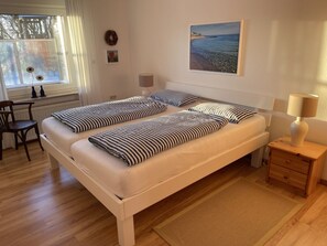 A double bed with striped bedding, a bedside table with a lamp, a chair, a window with a view of the sea, and a painting on the wall. - Holiday Apartment With 2 Bedrooms (Fehmarn)