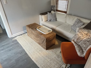 Ein Wohnzimmer mit einer Couch, einem kleinen Hocker mit Tablett, einer Lampe und einem Fenster mit Jalousien.