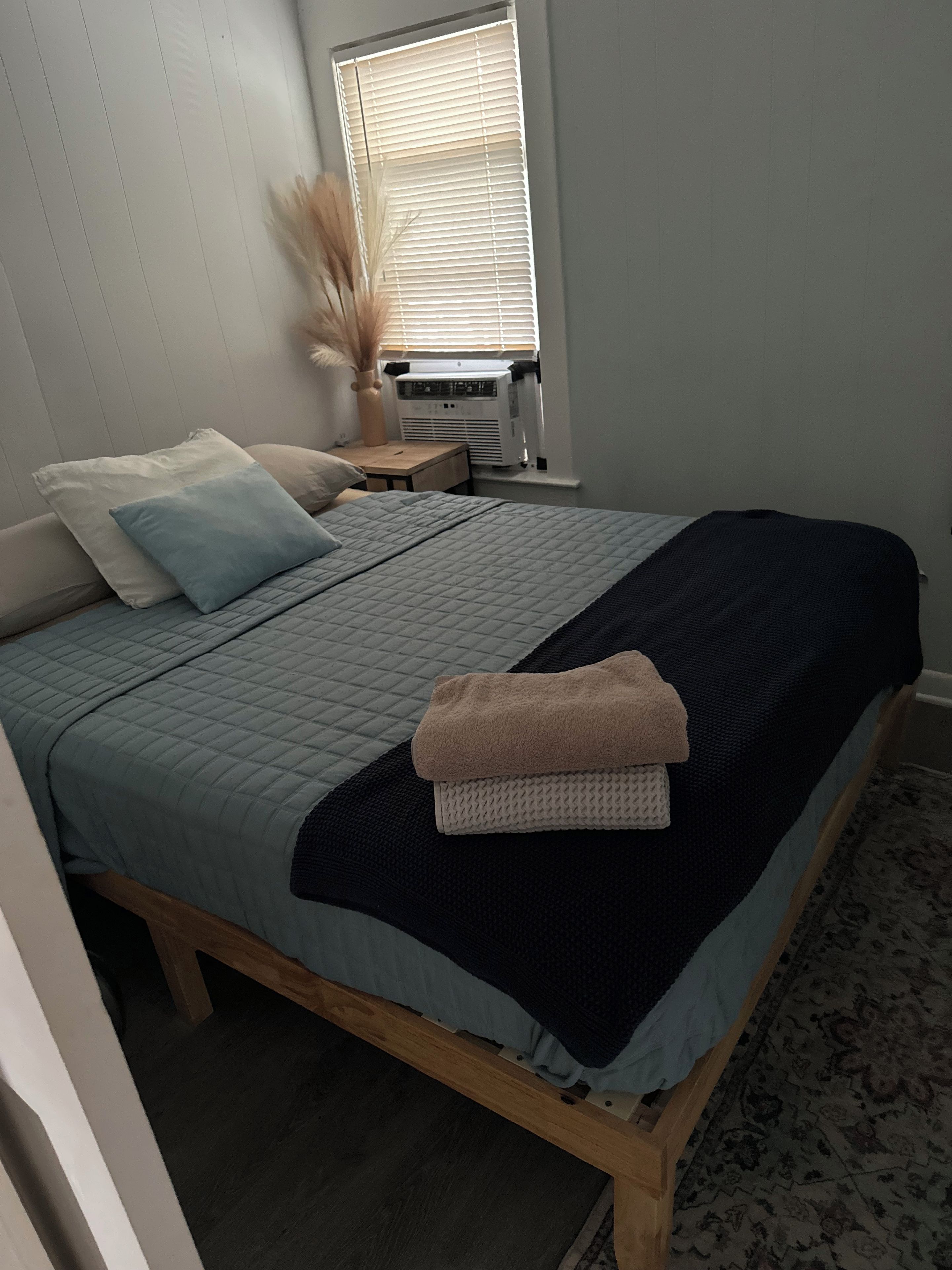 A bedroom with a bed, pillows, a bedside table with a lamp, and a window with blinds.
