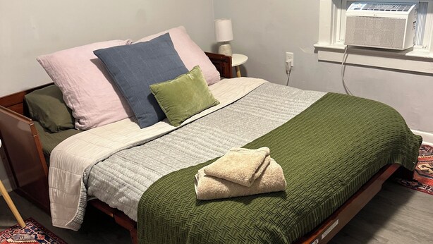 A neatly made bed with a green blanket and pillows, a patterned rug, a bedside table with a lamp, and a window with blinds. - Welcoming 3-bedroom house in Asbury Park Close to Downtown & Beach (Asbury Park)