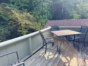 Eine Holzterrasse mit Tisch und Stühlen, umgeben von einem weißen Geländer, mit Blick auf einen bewaldeten Hang.
