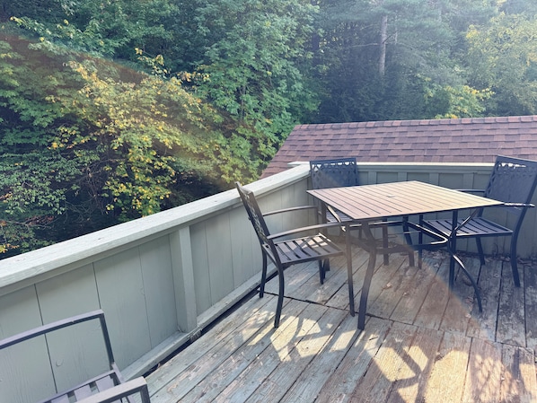 Une terrasse en bois, agrémentée d’une table et de chaises, entourée d’une balustrade blanche, et offrant une vue sur une colline boisée.