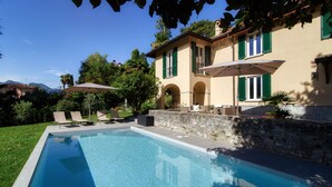 Une piscine avec des chaises longues et des parasols, un mur en pierre et un bâtiment aux volets verts.